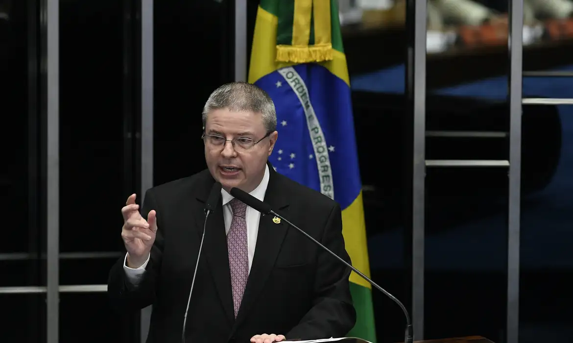 Antonio Anastasia em pé, falando em microfone e gesticulando com bandeira do Brasil ao fundo