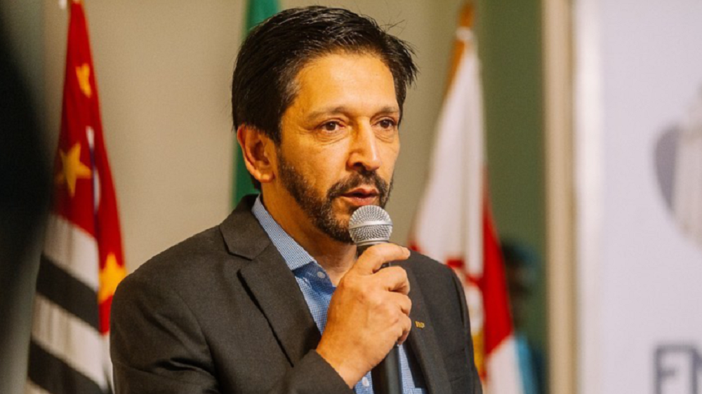 Ricardo Nunes falando em microfone com expressão séria, com bandeira do Brasil ao fundo