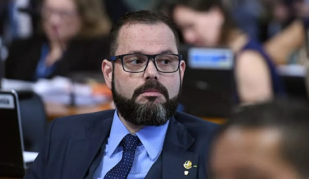 Jorge Seif olhando para o lado com expressão séria, de óculos de grau e roupa social em tons de azul
