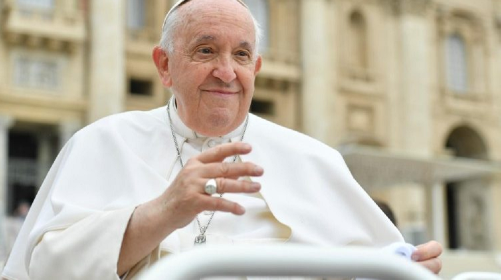 Papa Francisco acenando e sorrindo de forma tímida, sem olhar para a câmera