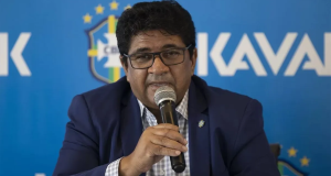 Ednaldo Rodrigues falando em microfone, de ôculos, com painel azul da CBF atrás