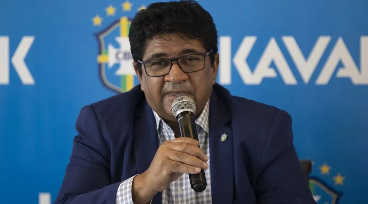Ednaldo Rodrigues falando em microfone, de ôculos, com painel azul da CBF atrás