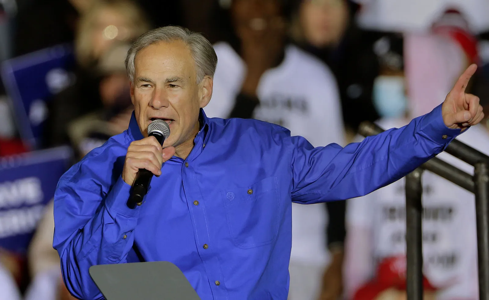 Greg Abbott de camisa social azul, falando em microfone e apontando para o lado