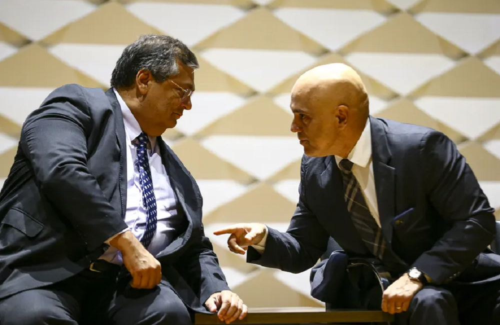 Flávio Dino e Alexandre de Moraes conversando, ambos sentados e próximos, de roupa social