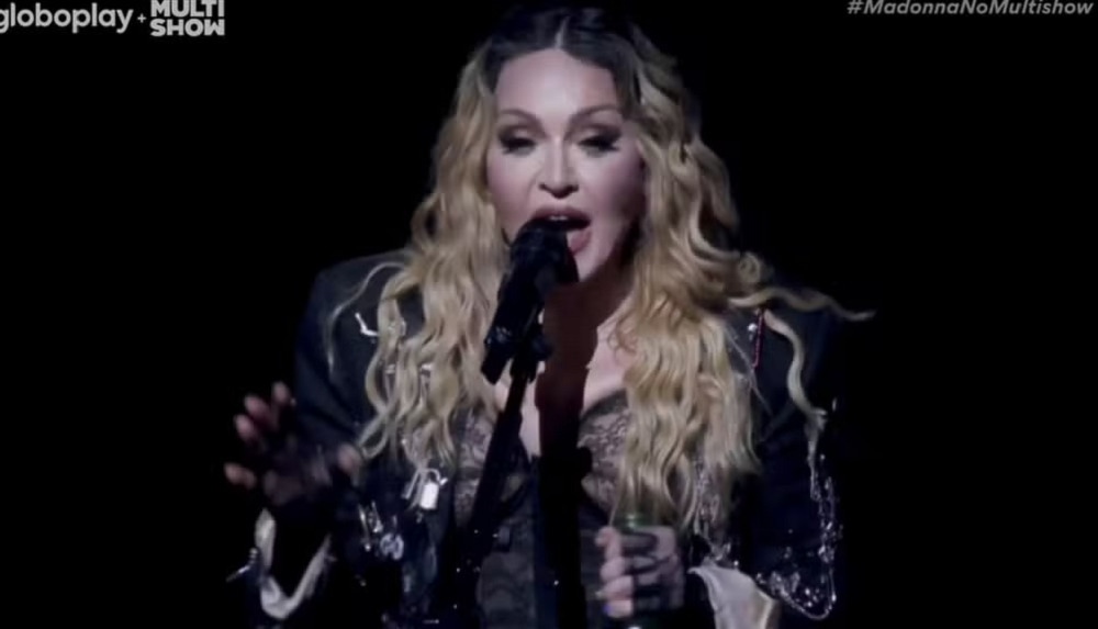 Madonna de roupa preta e cabelo solto, cantando em microfone