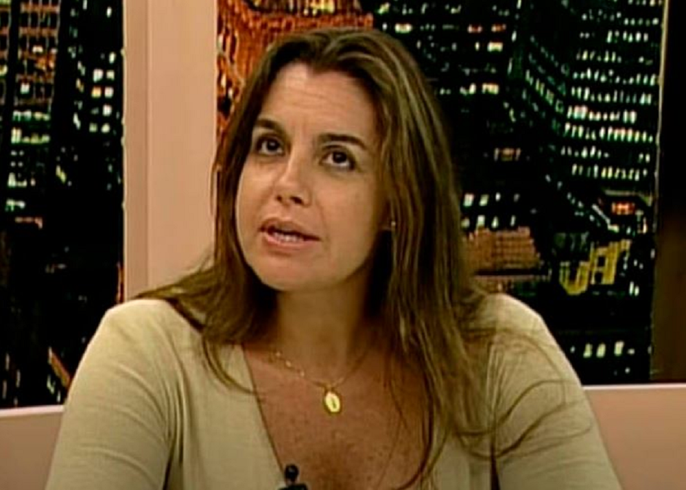 Maria Christina de cabelo solto e blusa bege, falando com expressão séria