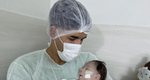 Zé Vaqueiro com touca e máscara, segurando Arthur no colo, em hospital