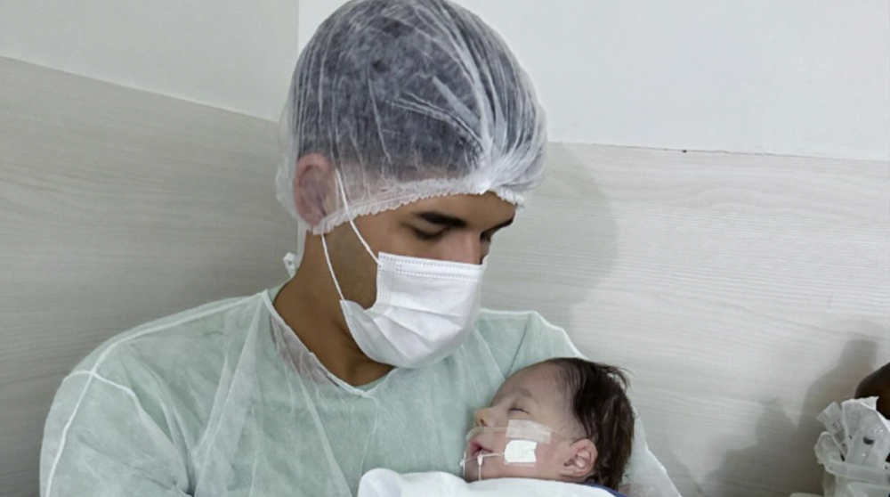 Zé Vaqueiro com touca e máscara, segurando Arthur no colo, em hospital