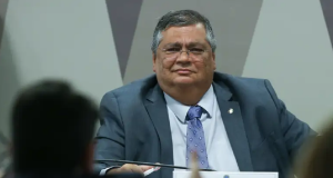Flávio Dino de óculos, de terno e gravata, olhando pra frente com expressão desconfiada