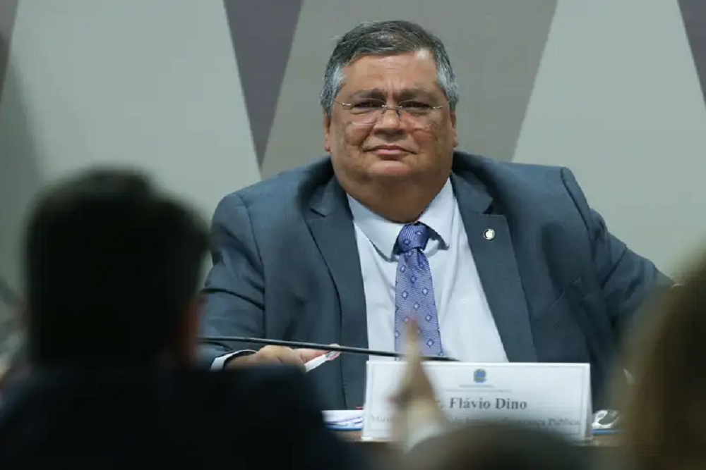 Flávio Dino de óculos, de terno e gravata, olhando pra frente com expressão desconfiada