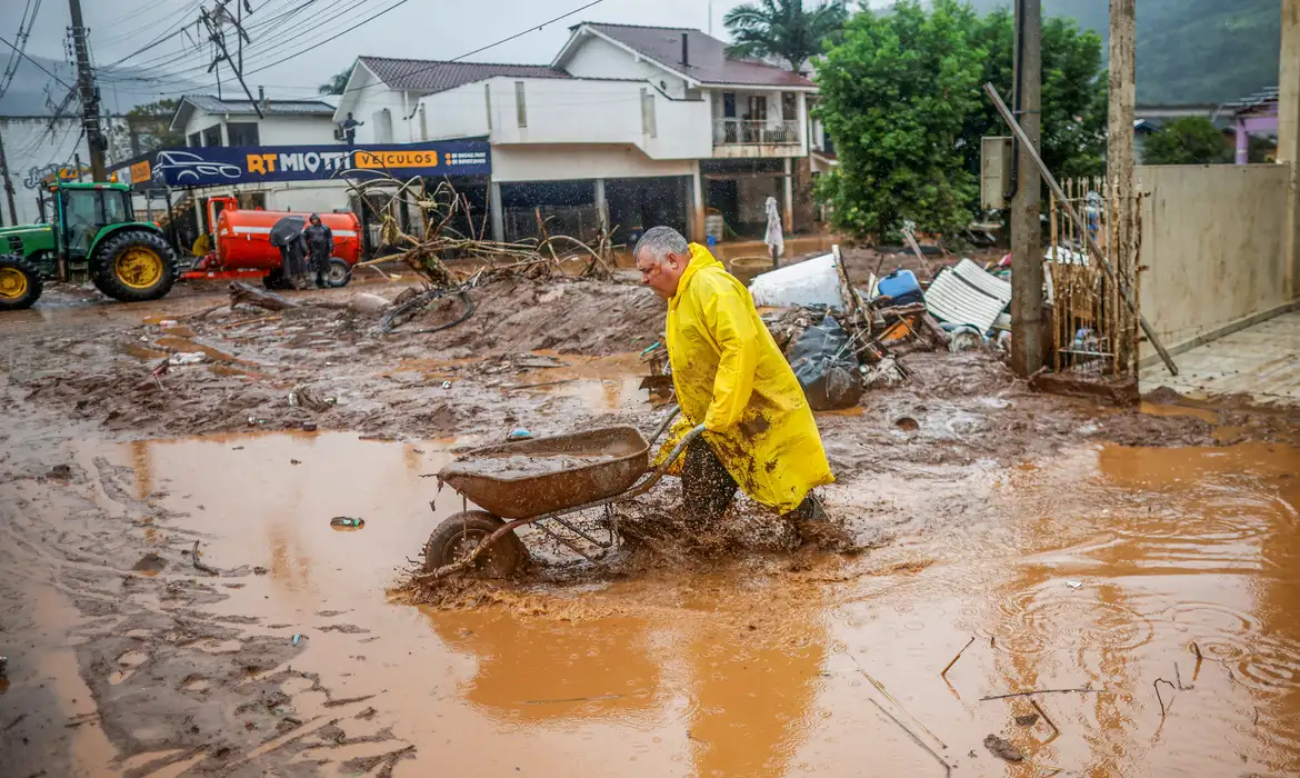Homem de capa de chuva amarela andando e empurrando carrinho de mão em rua alagada