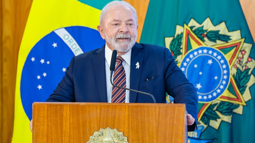 Lula sorrindo, de terno e gravata, falando em microfone com bandeiras ao fundo