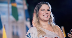 Marília Mendonça sorrindo, de roupa estampada, segurando microfone, de cabelo solto, sem olhar pra câmera