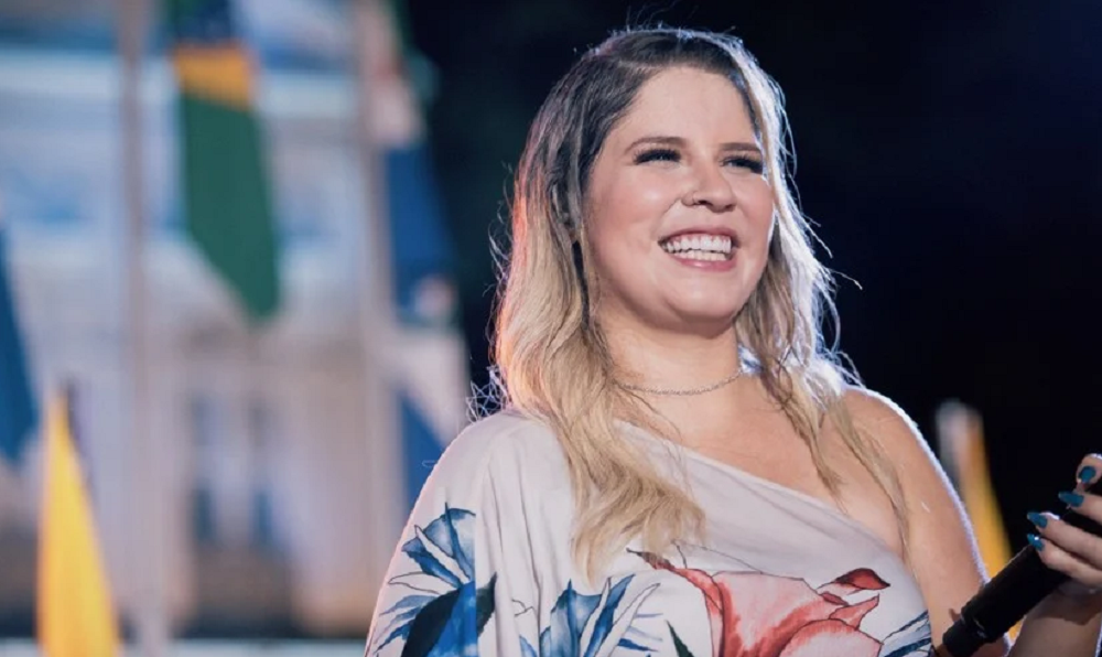 Marília Mendonça sorrindo, de roupa estampada, segurando microfone, de cabelo solto, sem olhar pra câmera