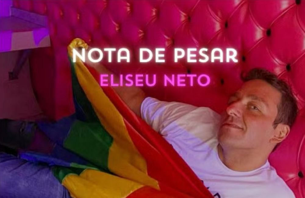 Eliseu Neto em local cor de rosa, de camiseta branca, segurando bandeira 