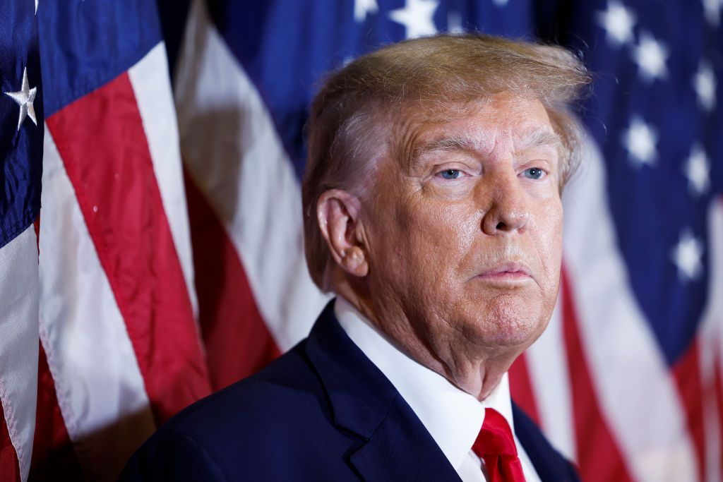 Donald Trump fazendo careta sem olhar para a câmera, com bandeiras dos Estados Unidos ao fundo