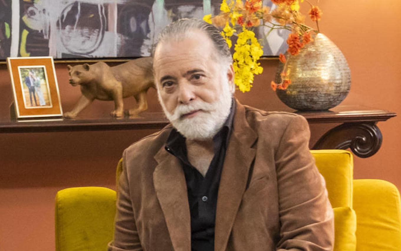 Tony Ramos de roupa marrom, sentado em poltrona amarela, sem sorrir, olhando pra câmera