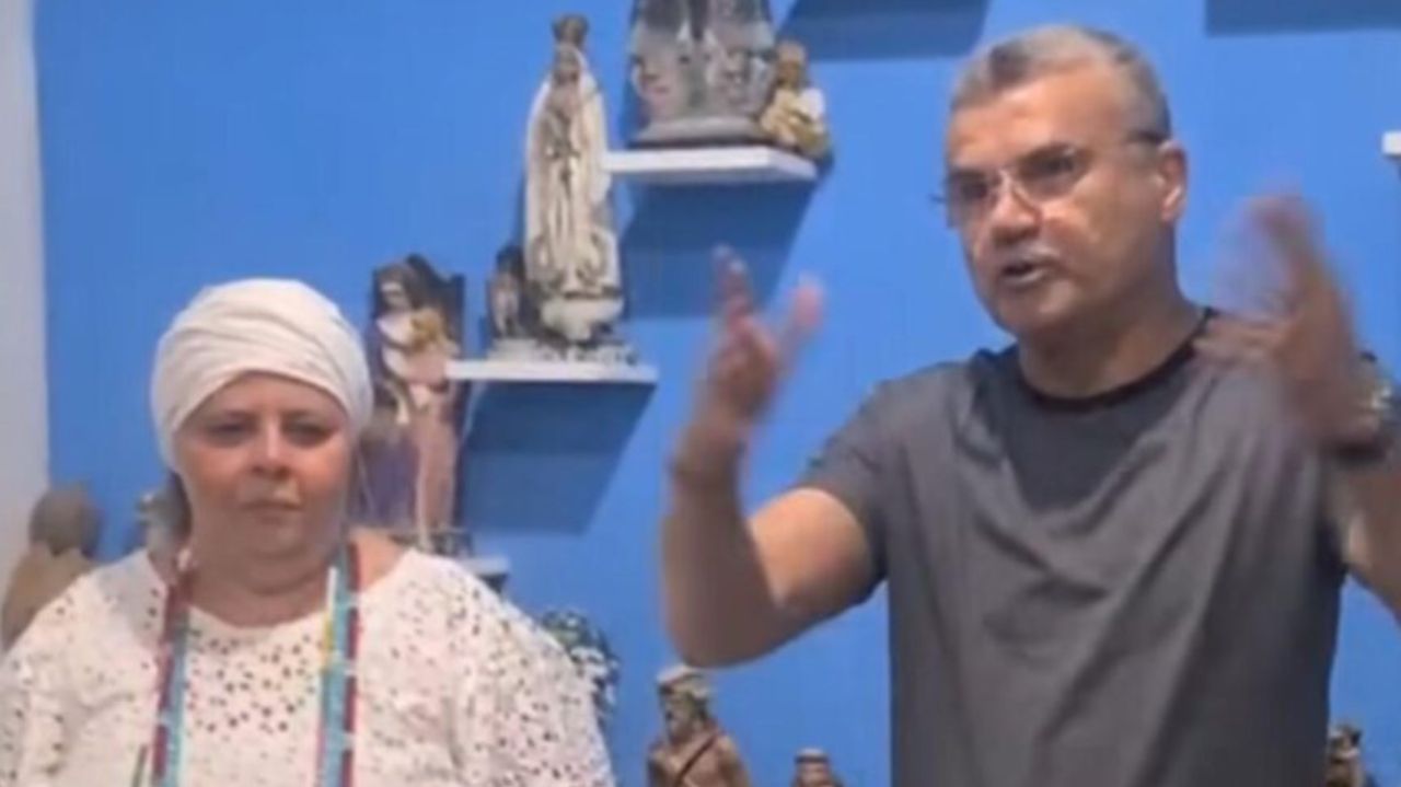 Mãe de santo ao lado de pastor, que está de camisa cinza, falando e gesticulando