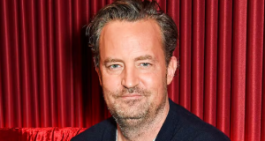 Matthew Perry olhando para a câmera sem sorrir, em foto feita em fundo vermelho