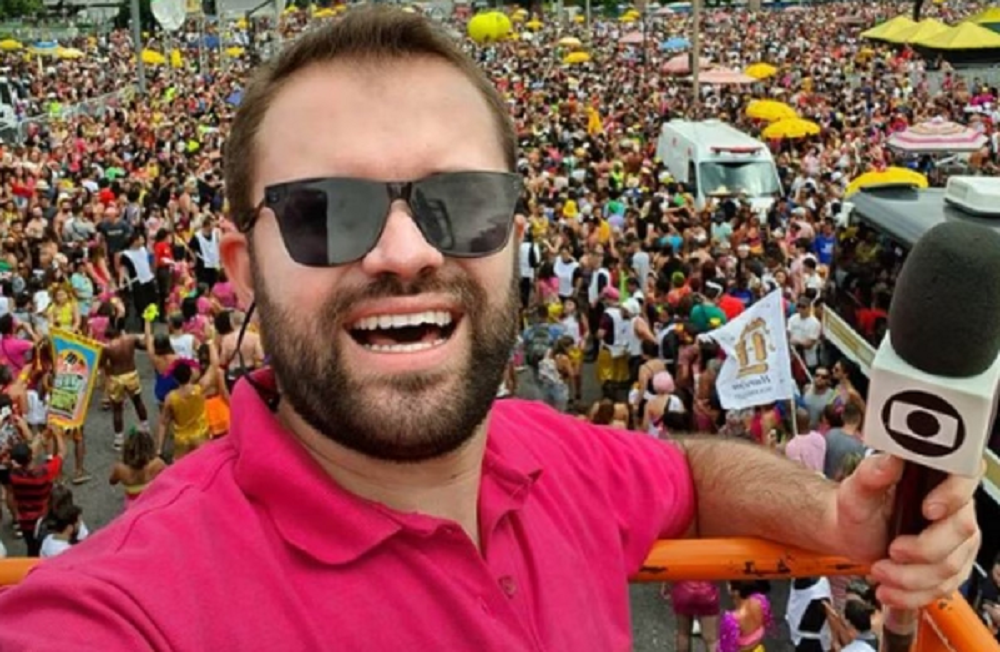 Aluísio Marques sorrindo de camisa rosa e óculos escuros, segurando microfone da Globo