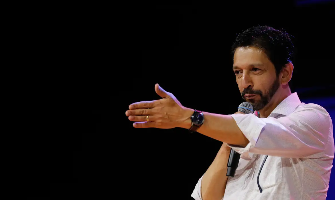 Ricardo Nunes de perfil, no canto direito da foto, falando em microfone e apontando pra frente