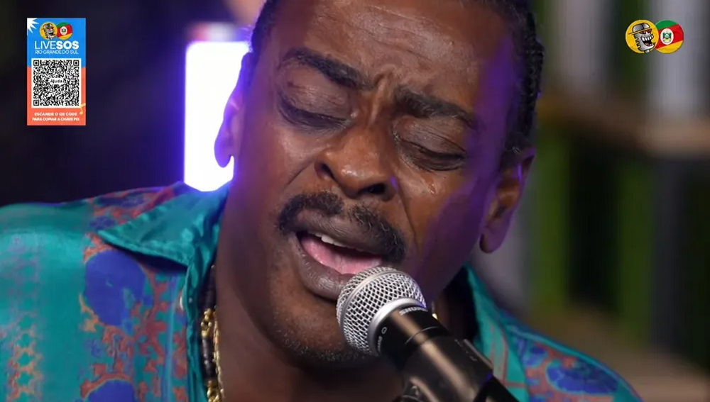 Seu Jorge de roupa estampada e olhos fechados, cantando em microfone, em close