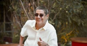 Zeca Pagodinho de camisa branca e óculos escuros, fazendo sinal de joia e sorrindo