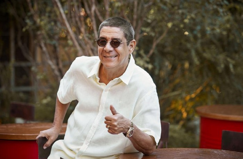 Zeca Pagodinho de camisa branca e óculos escuros, fazendo sinal de joia e sorrindo