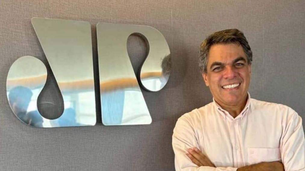 André Ramos sorrindo, de camisa branca e braços cruzados, perto de símbolo prateado da Jovem Pan