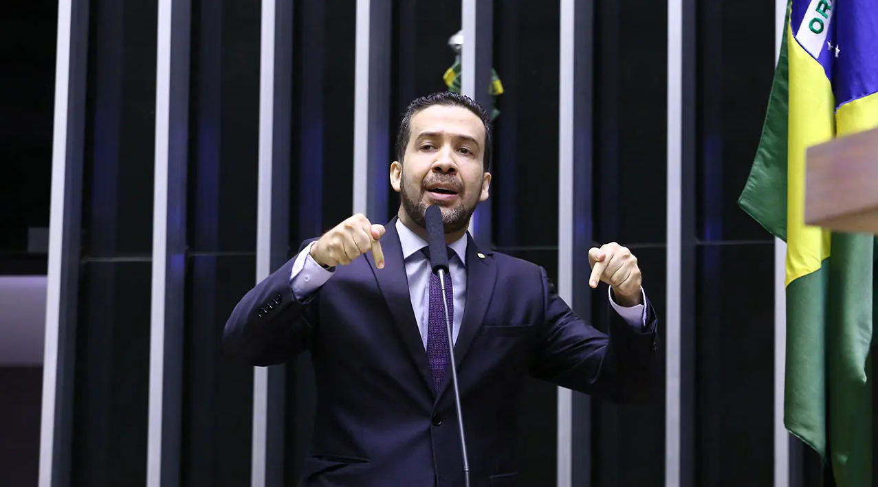 André Janones com expressão séria, falando em microfone no plenário e apontando para baixo