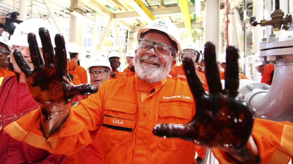 Lula de roupa laranja sorrindo e mostrando mãos sujas de petróleo para a câmera