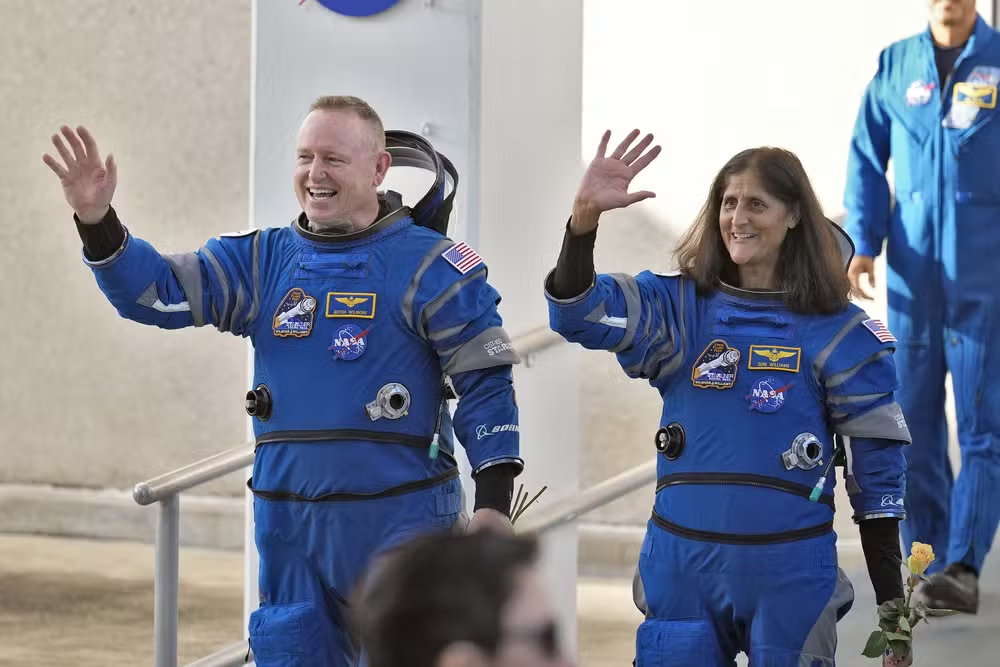 Butch Wilmore e Suni Williams, astronautas da Nasa, de uniforme, acenando e sorrindo sem olhar pra câmera