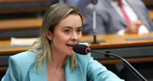 Deputada federal Júlia Zanatta de roupa azul e cabelo solto, falando em microfone sem olhar pra câmera
