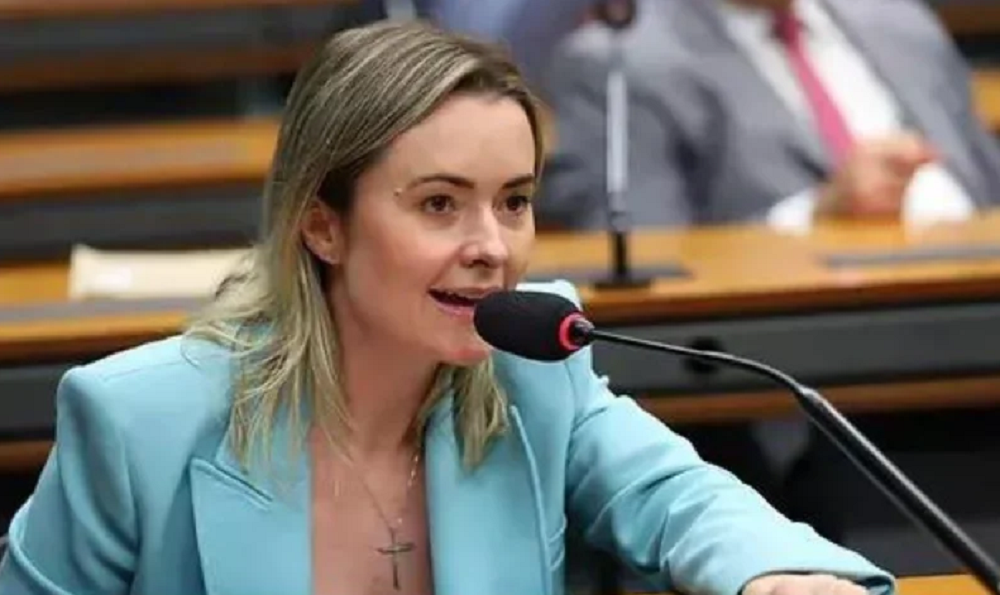 Deputada federal Júlia Zanatta de roupa azul e cabelo solto, falando em microfone sem olhar pra câmera