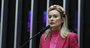 Deputada federal Julia Zanatta de roupa cor de rosa, segurando celular, com expressão séria e flores na cabeça