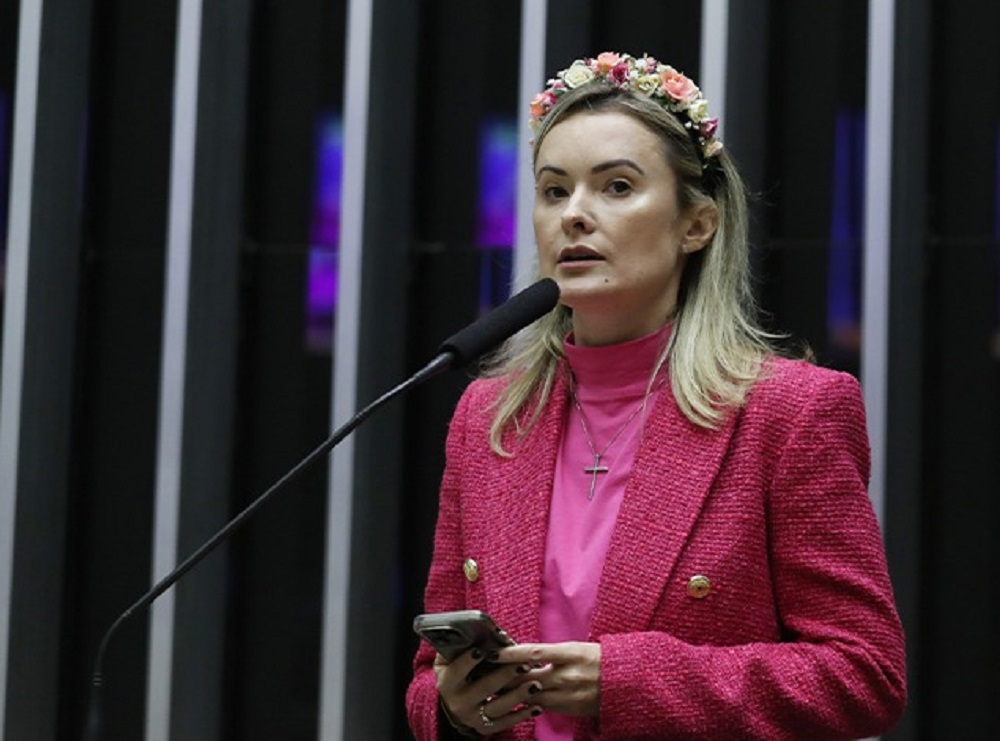 Deputada federal Julia Zanatta de roupa cor de rosa, segurando celular, com expressão séria e flores na cabeça