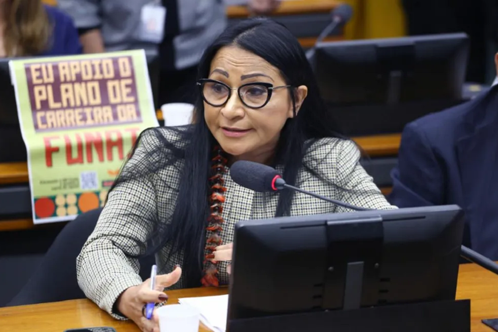 Deputada federal Silvia Waiãpi falando em microfone com expressão séria, de óculos e cabelo solto, sentada