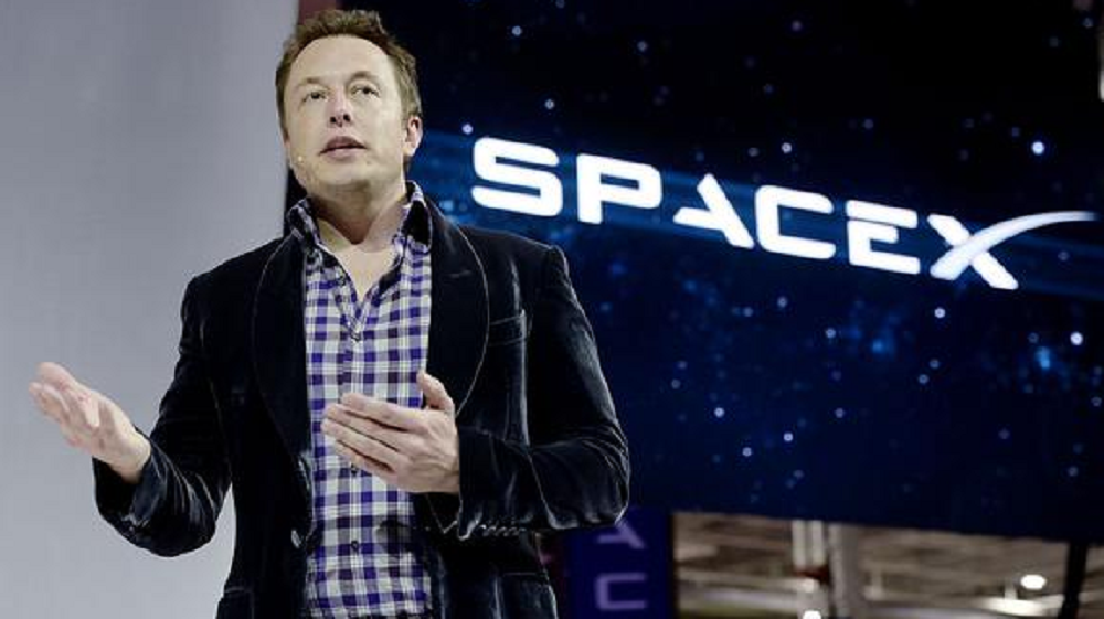 Elon Musk de roupa xadrez, falando e gesticulando com telão da SpaceX atrás