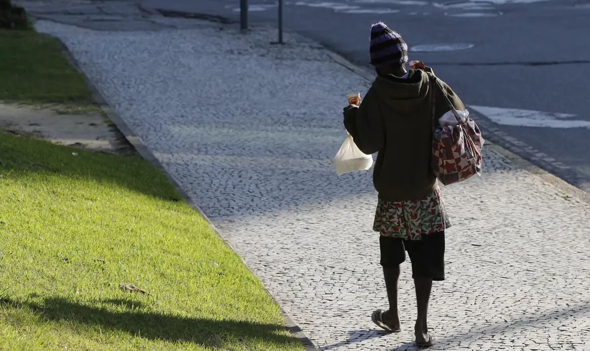 Pessoa em situação de rua carregando marmita e caminhando, de costas