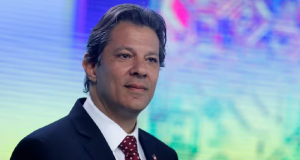 Fernando Haddad com expressão distraída, sem olhar para a câmera, perto de microfones e de painel colorido