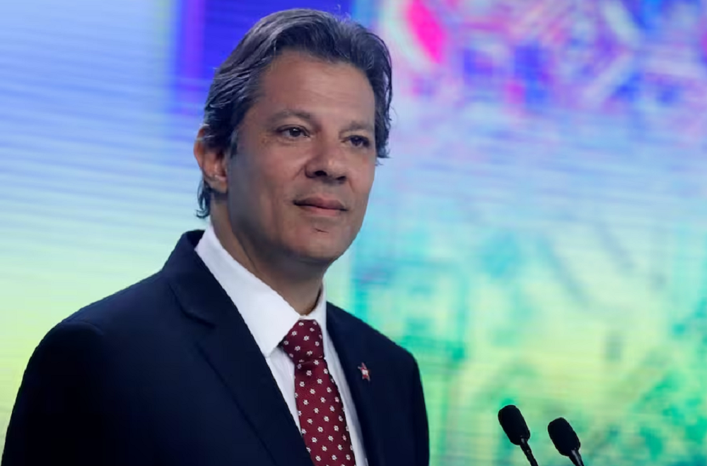 Fernando Haddad com expressão distraída, sem olhar para a câmera, perto de microfones e de painel colorido