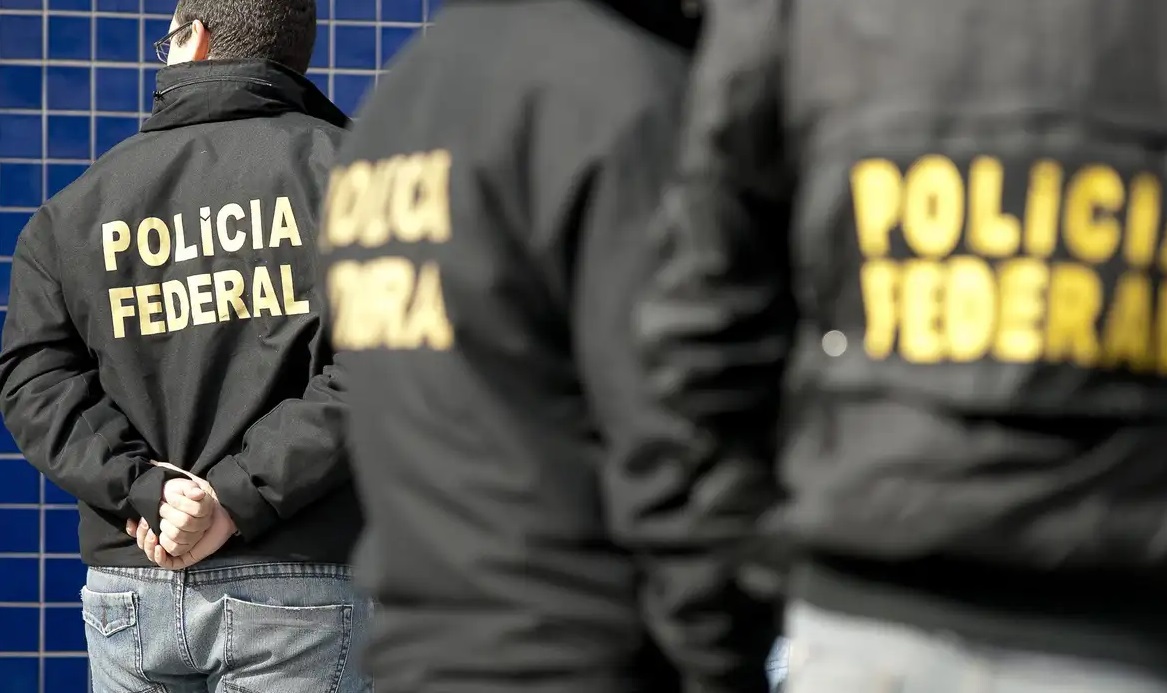 Três agentes da Polícia Federal uniformizados, de costas, lado a lado