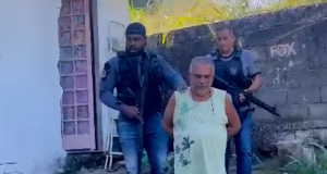 Pai de santo com as mãos pra trás sendo conduzido por dois agentes