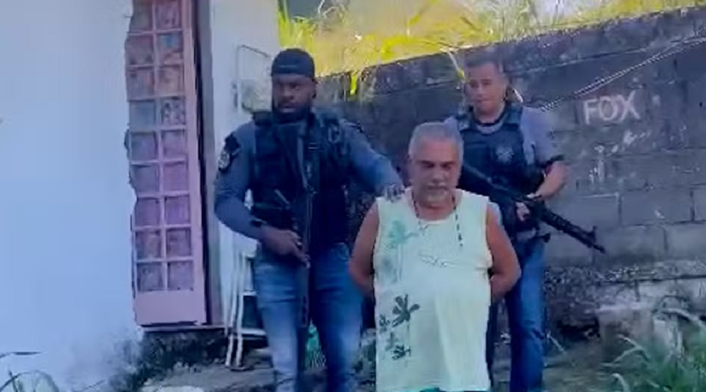 Pai de santo com as mãos pra trás sendo conduzido por dois agentes