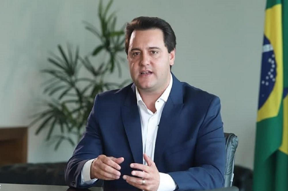 Ratinho Jr. falando e gesticulando com expressão séria e bandeira do Brasil ao fundo
