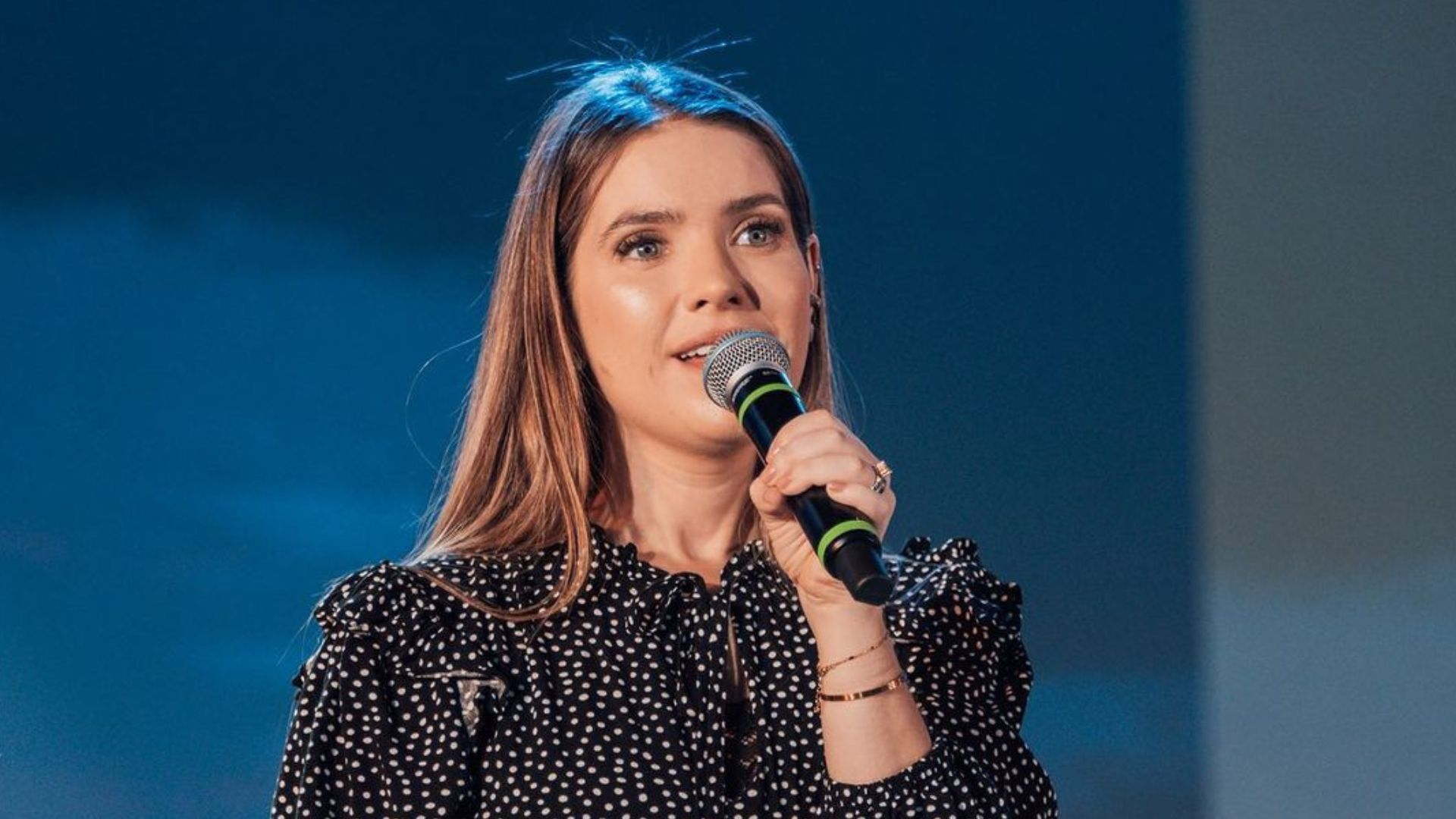 Camila Campos de roupa de poá preta e branca e cabelo solto, cantando em microfone