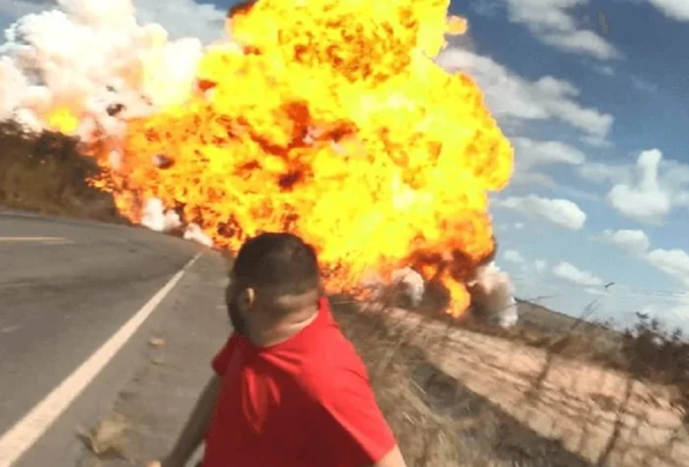 Homem de camisa vermelha correndo e olhando para trás, caminhão pegando fogo ao fundo