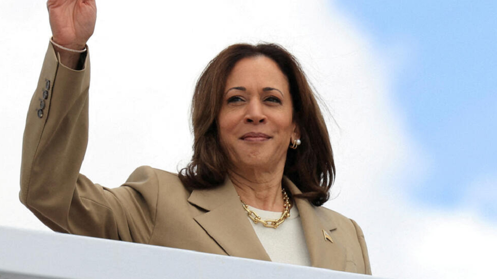A vice-presidente dos Estados Unidos, Kamala Harris, de roupa bege, com sorriso tímido, acenando, com expressão esperançosa