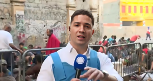 Marcos Guimarães falando em microfone da Record e olhando para a câmera, com camisa social branca e colete à prova de balas