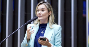A deputada federal Júlia Zanatta (PL-SC) de roupa social azul, falando em microfone e segurando celular, de cabelo solto